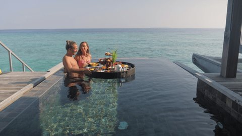 Beautiful young couple eat breakfast in private pool from floating tray. Couple enjoying tropical vacation in overwater bungalow  