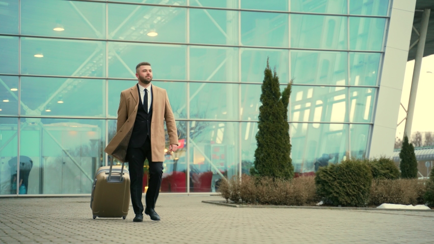 young bearded male tourist in a business suit and coat goes from the airport with a suitcase arrival or departure. on the street background of a modern building. Stylish Businessman walking city trip Royalty-Free Stock Footage #1069352269