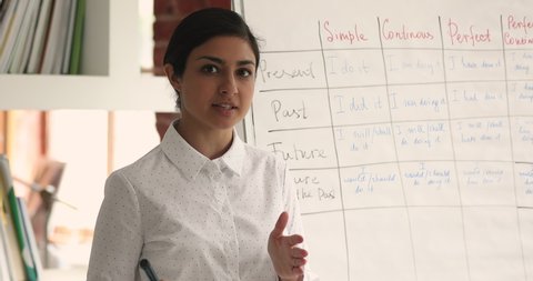 Indian mixed race teacher standing near flip chart in classroom looking at camera telling about foreign language tenses, teaches English grammar using video call. Remote education and tutoring concept