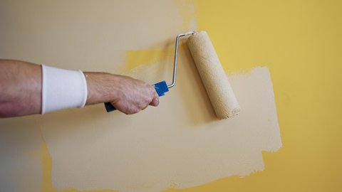 Roller painting yellow wall with creamy paint. Hand of a worker doing renovation the room with light color. Close-up. Makeover concept.