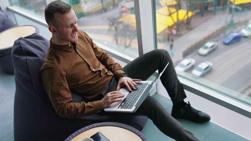 Handsome businessman in office by the window. Young man in brown shirt and trousers sitting on a comfortable ottoman and working on a laptop. City view background. | Shutterstock HD Video #1069357807