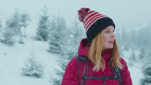 Happy portrait of blonde woman wearing winter outfit watching a snowy landscape and looking at the camera while snow falling. Winter holiday concept.