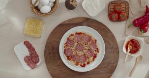 Preparing Italian salami pizza by a recipe from the online culinary course, ingredients for cooking. Woman hands taking plate with uncooked salami pizza from the table.