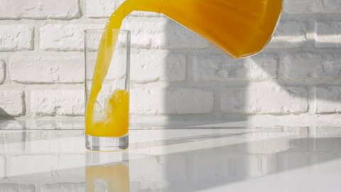 Pour yellow lemonade into a glass from a jug against a white brick wall background. Fresh orange juice. A glass with a fruity orange drink on a white kitchen table.