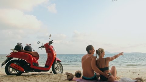 Scooter road trip. Lovely couple on red motorbike in white clothes on sand beach. People walking near the tropical palm trees, sea. Motorcycle rent.