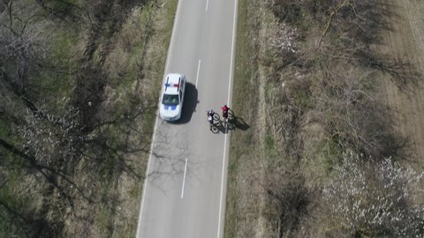 Aerial view of a young couple of cyclists riding on an asphalt road driving towards a police car between a field and a forest on a beautiful spring day.