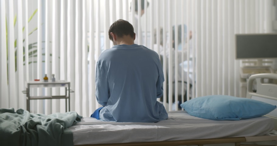 Back view of young desperate man sitting at hospital bed alone. Sick male patient sitting on bed at clinic upset for serious disease diagnosis feeling worried | Shutterstock HD Video #1069361842