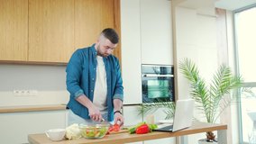 young handsome bearded man preparing food online at home in the kitchen using laptop. Male cuts vegetables at the table for a vegetarian salad and watches instructional video tutorials cooking recipe