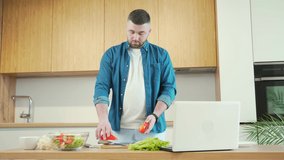 young handsome bearded man preparing food online at home in the kitchen using laptop. Male cuts vegetables at the table for a vegetarian salad and watches instructional video tutorials cooking recipe