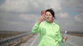 Mature woman walks along the road and talks on a video call using a phone and headphones. People in nature. Talk to family and friends over a walk.
