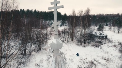 Aerial view through the cross on the dome of an old wooden Christian church located in a remote abandoned village. In the distance, a pilgrim wanders through snowdrifts and a snow-covered forest.