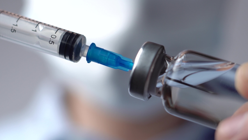 Doctor in a white medical mask and blue gloves holds in his hands a syringe and a glass vaccine bottle at the hospital. Physician dials the medicine into the syringe | Shutterstock HD Video #1069372387