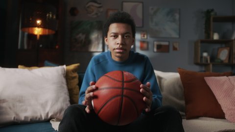 Serious black man teenager holding basketball ball and sitting in living room, young teen athlete, sport portrait at home, kid preparing for watching professional game on tv.