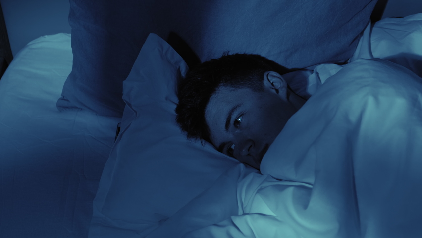 Night insomnia. Sleep disorder. Depression anxiety. Disturbed thoughtful guy awake late lying in bed alone at home in dark blue light. | Shutterstock HD Video #1069375492