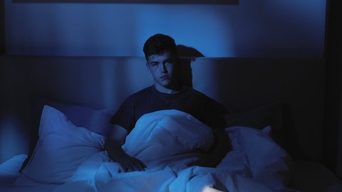 Night insomnia. Sleep disorder. Depression anxiety. Tired bored pensive guy cant rest awake late yawning sitting alone in bed at home in dark blue light.