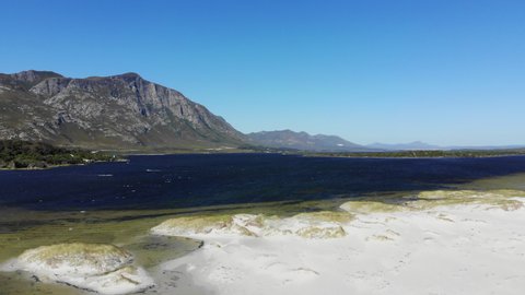 Scenic drone footage of kiteboarding in the lagoon with mountains in the background in Hermanus in South Africa. Drone flying backwards away from the kites and over a sand dunes and beach.