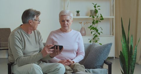 Caucasian couple of pensioners old family grandparents husband and wife sitting on couch at home looking at screen of mobile phone arguing quarreling over new technologies receive bad message online