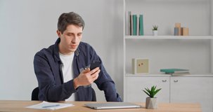 Caucasian millennial business man adult young guy entrepreneur sitting in office home holding phone in hand talking into webcam smartphone communicating using remote video chat conference mobile app