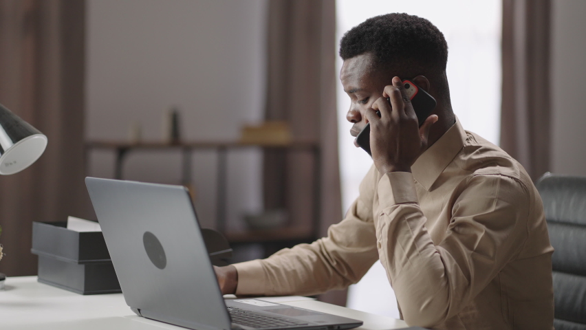 Black man entrepreneur is calling by phone and making notes, sitting in front of laptop, working from his home office, negotiation by cellphone | Shutterstock HD Video #1069383013