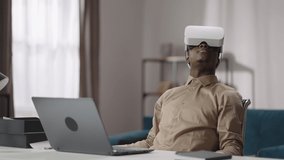 young black man is using head-mounted display for playing video games, sitting at table with laptop and looking around, virtual reality technology