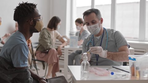 Waist up of grey-haired male Caucasian doctor wearing medical mask and gloves, sitting by desk in clinic, disinfecting skin on shoulder of young man, then giving him vaccine injection