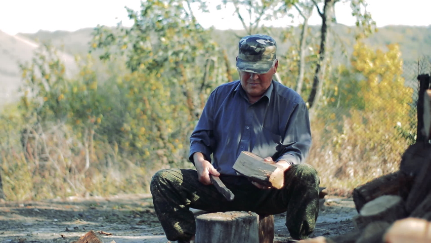 Old man chop wood sitting. View from the front. Outdoors in the daytime.