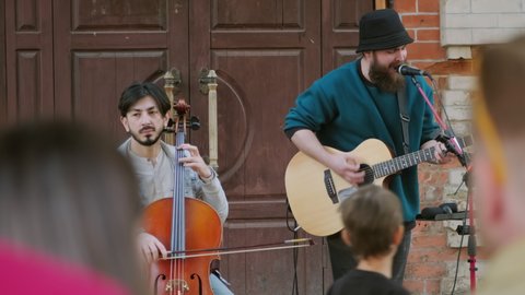 Medium shot of street musicians playing cello and acoustic guitar while performing song for crowd outside