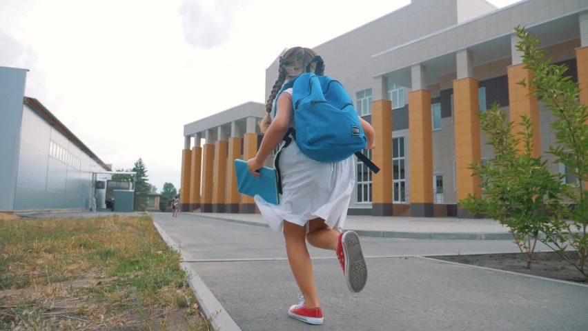 A small schoolgirl with a backpack and a book runs through the Park to school. A girl with a school backpack hurries to class. Educated children. | Shutterstock HD Video #1069389538