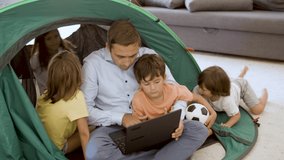 Father and kids playing camping at home, using laptop, learning how to set tent. Childhood or parenthood concept