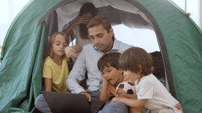 Dad and kids playing camping at home, using laptop for watching movie, browsing internet or video call from tent set in living room. Childhood or parenthood concept