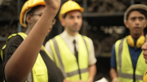 Close up of black woman foreman leader wearing uniform protection for safety holding black clipboard discussing with worker team checking stock.