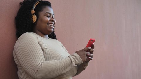 Happy Afro woman messaging online with mobile smartphone while listening music with wireless headphones