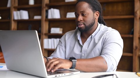 Confident African American businessman wearing shirt using laptop in modern office, checking the time from a watch on his wrist, in a hurry to finish work, has a deadline with online work