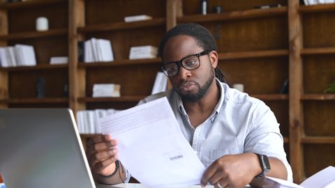 Serious African-American businessman looking through mailing correspondence, concentrated man opens envelope and take letter out, reading a document, received an important notification by mail
