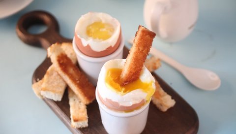boiled egg in eggcup on wooden board with crispy toast. Woman hand dipping toast into a egg.
