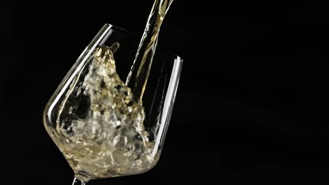 Pouring gold wine into goblet at black background. Close-up of filling wine glass with white wine in super slow motion. White wine forms beautiful wave in glass with copy space