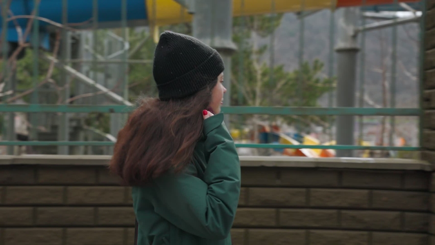 A young woman in a jacket and hat walking along the street along the fence. Side view