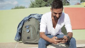Young lonely traveller blogger sitting on bench with his backpack next to him chatting with his fan followers on social networks laughing and typing text on the phone. New technology of communication