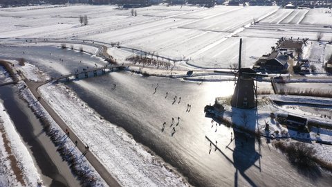 Aerial view of beautiful Winter landscape with frozen canals and snowy meadows, people ice skate past historic windmills in the Netherlands.
