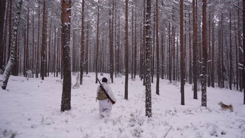 A hunter in camouflage clothing walks through the winter forest with a dog. A man removes a gun from his shoulder, preparing to shoot