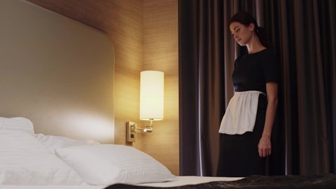 young, beautiful maid in uniform and apron making bed in hotel room