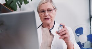 Senior female pharmacy doctor woman in lab coat offers vaccine flask to remote client online using laptop webcam chat.