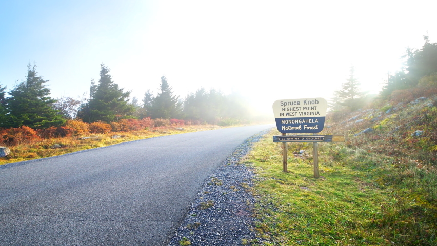 Heavy fog morning mist floating above road to Spruce Knob mountain ridge overlook in Allegheny mountains with sign for Monongahela National forest at sunrise with sun sunlight sunburst, West Virginia
