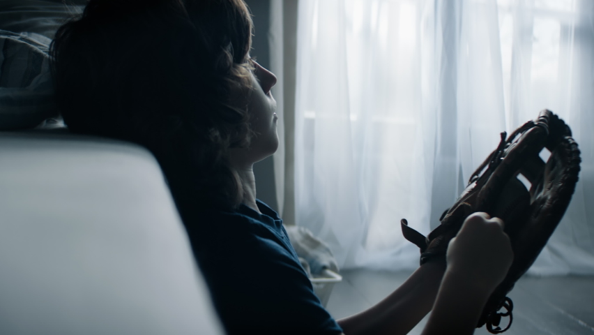 Silhouette portrait of cute little Caucasian kid sitting in his bedroom, playing baseball catch with himself at home. Shot with 2x anamorphic lens