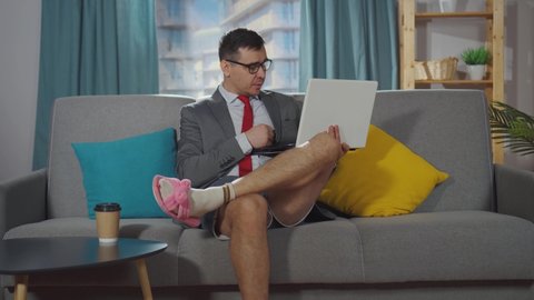 Serious office worker in a suit and shorts sits in the living room and talks on a laptop at work