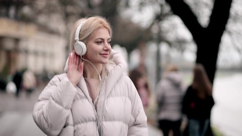 Slow motion of happy young woman in headphones dancing outdoors n the city park having fun alone. Joyful attractive blonde carefree woman listening to music with smartphoneの動画素材