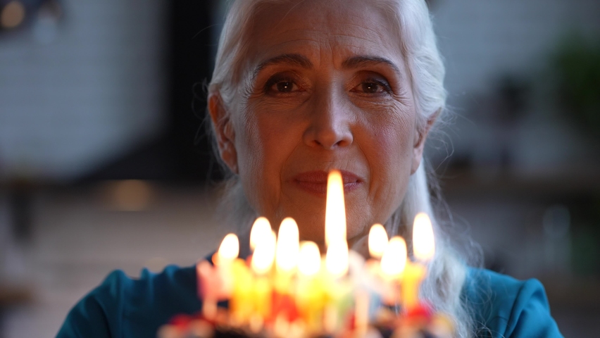 Close-up of attractive cheerful elderly woman with white hair blowing candles on colored Happy birthday lettering during anniversary celebration. Portrait of happy gray-haired female during holiday | Shutterstock HD Video #1069440553
