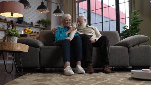 Cheerful white-haired aged people enjoying domestic leisure sitting on sofa with cute puppy while robot vacuum cleaner working. Modern elders spending free time while robotic cleaner cleaning carpet Stockvideó