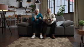 Advanced gambling old people with gray hair playing video games using joysticks while sitting together on couch. Young at heart modern elderly family enjoying domestic gaming activity during free time