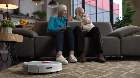 Close-up of robotic cleaner washing carpet while advanced old people with white hair enjoying free time using tablet and communicating with little chihuahua dog. Modern aged family during home leisure